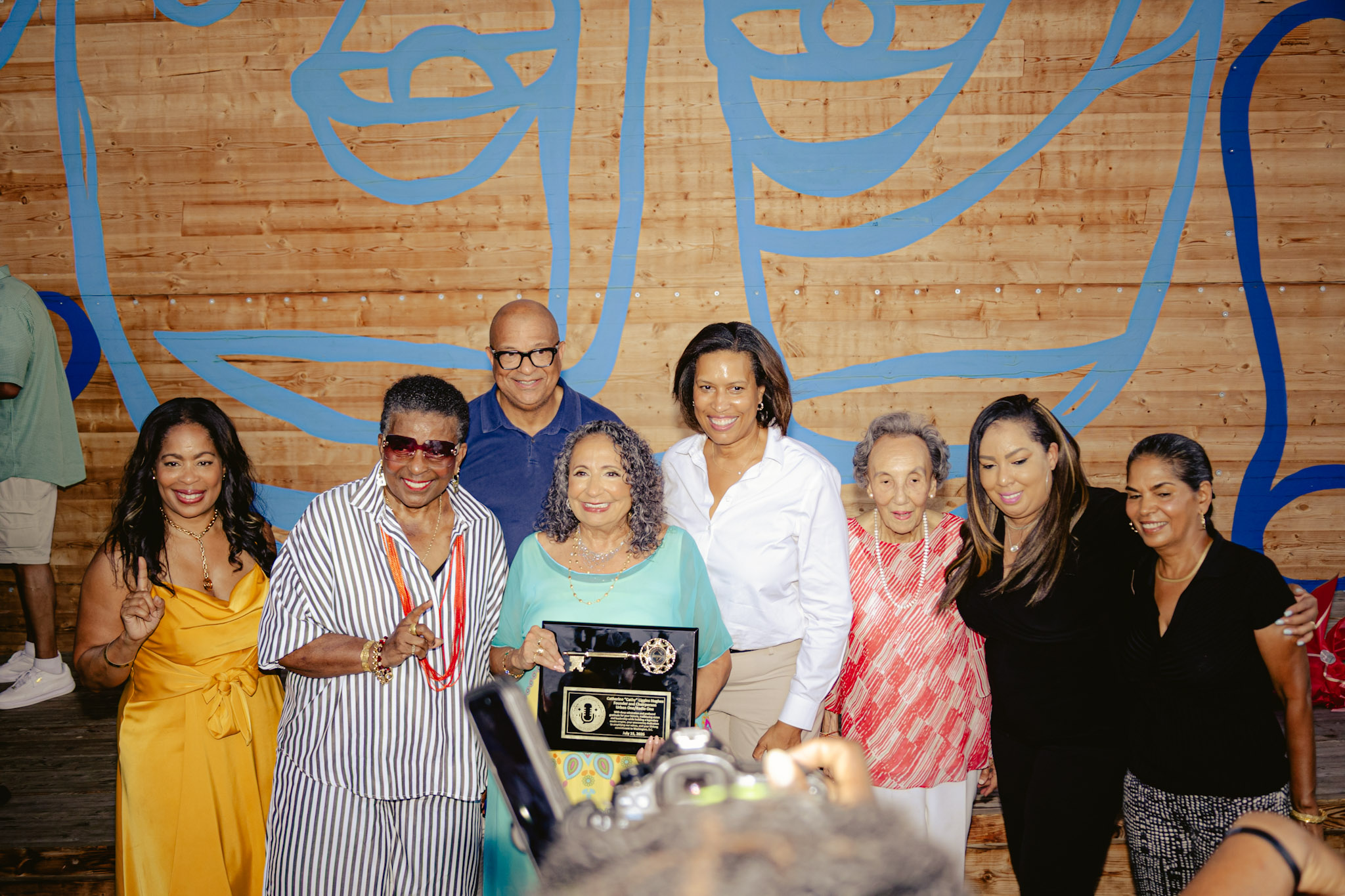A group of diverse people posing together, holding an award plaque. The background features a colorful mural on a wooden wall.