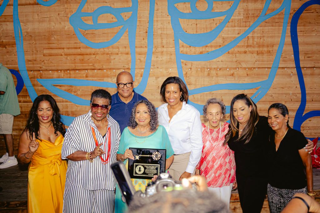 A group of diverse people posing together, holding an award plaque. The background features a colorful mural on a wooden wall.